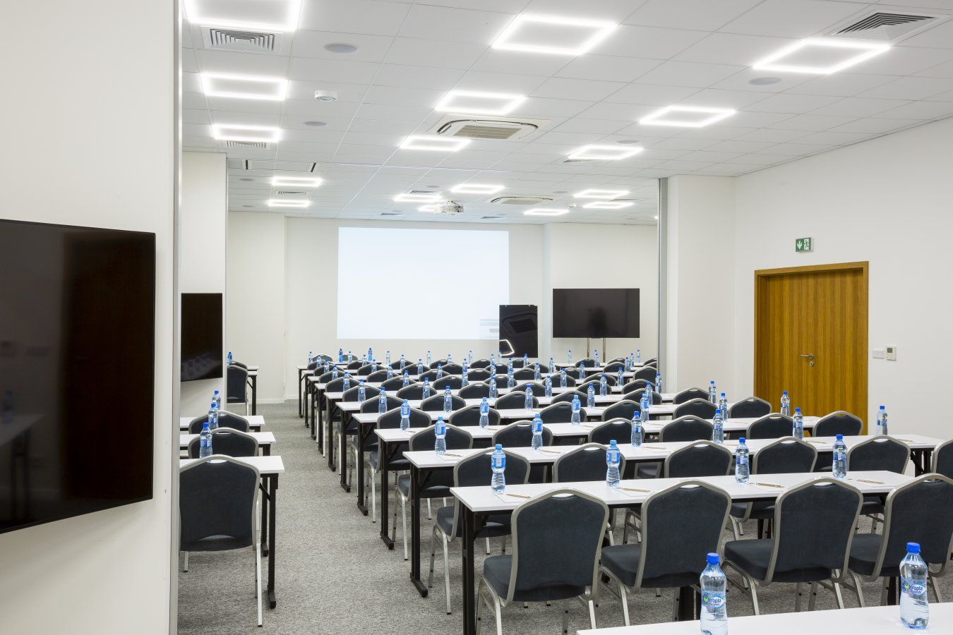 Uma sala de conferências com mesas, cadeiras e garrafas de água dispostas em fileiras, equipada com uma tela de projetor e uma televisão de tela plana grande.
