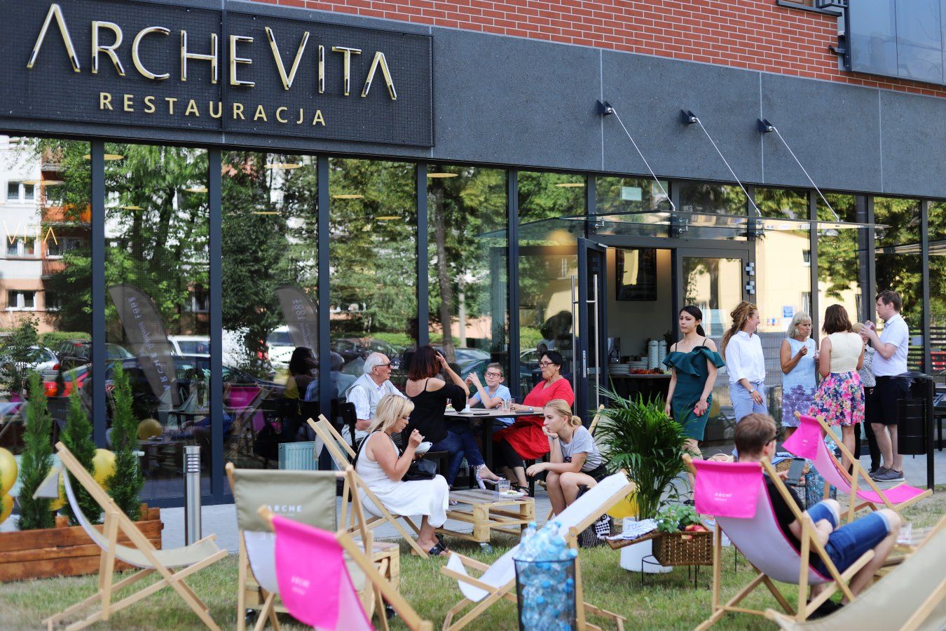 Un espace extérieur aménagé avec des tables et des chaises, une enseigne indiquant « ARCHEVITA RESTAURACJA », et un bâtiment doté de grandes fenêtres.