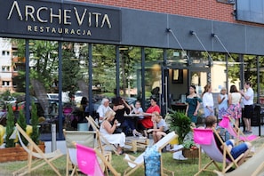 An outdoor seating area with tables and chairs, a sign for 'ARCHEVITA RESTAURACJA', and a building with large windows. - Arche Residence Lodz (Łódź)