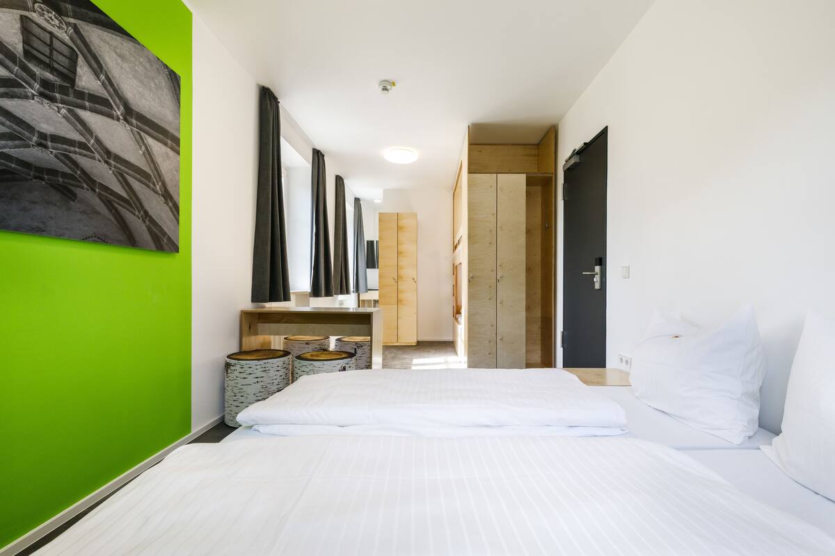Une chambre d’hôtel avec un grand lit, un mur d’accent vert, une armoire en bois, un miroir et un petit coin salon avec une table et des chaises.