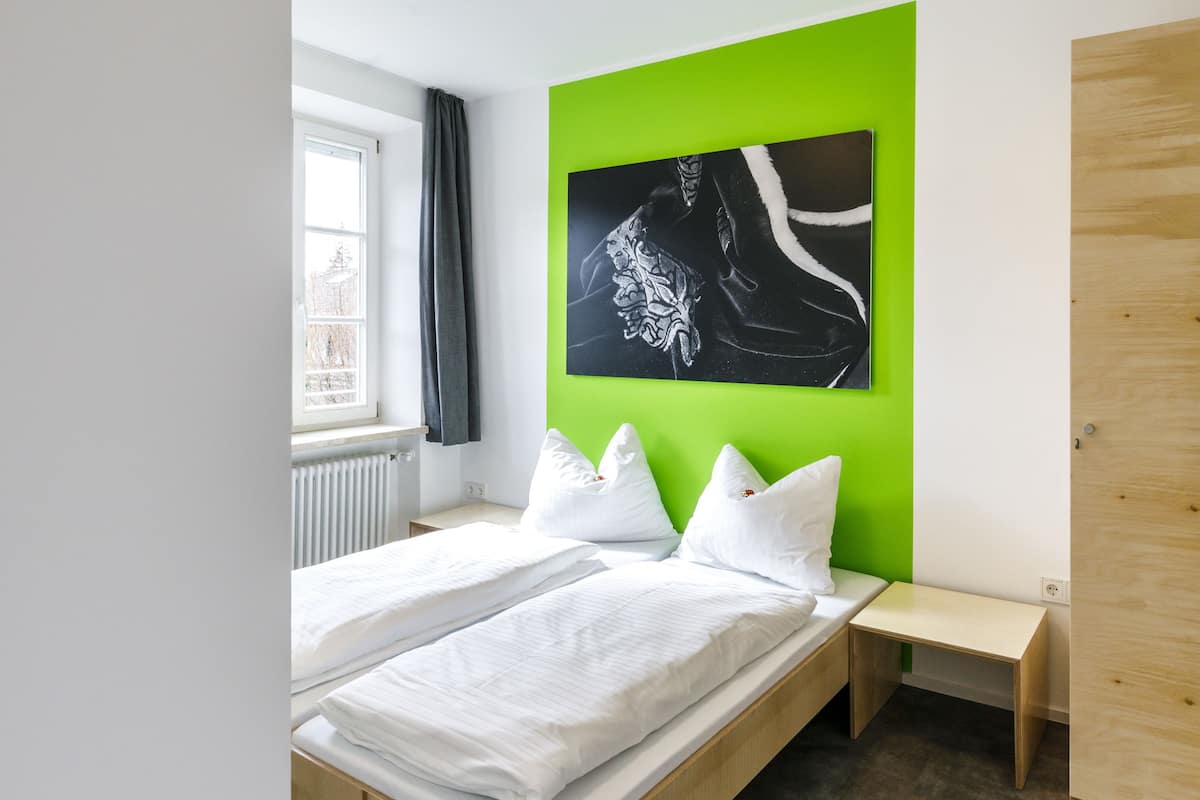 Une chambre moderne avec un grand lit, une table de chevet, un banc, un mur d’accent vert et une œuvre d’art en noir et blanc.