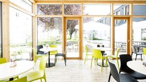 Een lichte, open eetruimte met grote ramen, een houten plafond en een mix van gele en zwarte stoelen rond witte tafels.