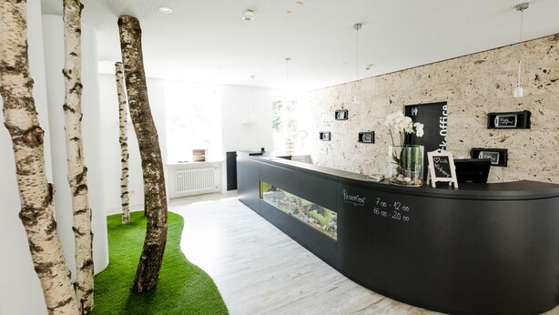 Een receptiezone met een modern design, herkenbaar aan kolomvormige structuren die doen denken aan berkbomen, een zwart receptiebalie en een uitzicht op de stad.