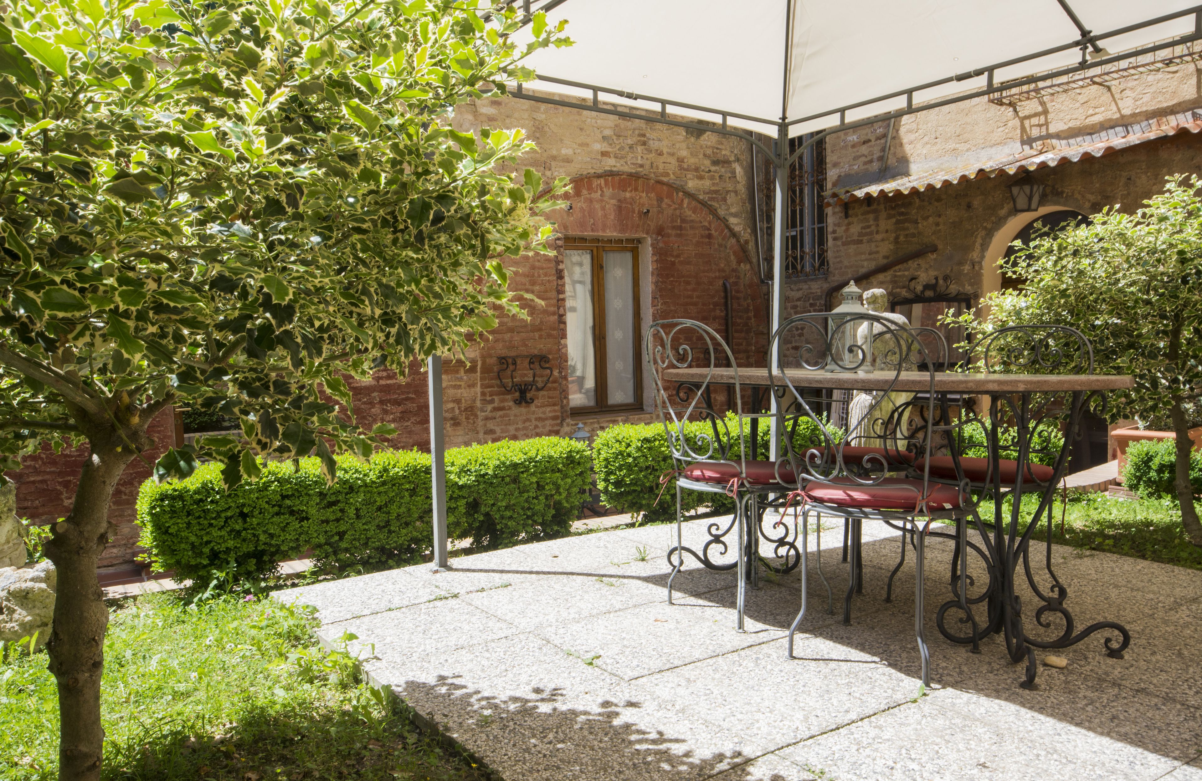 An outdoor patio with a metal table and chairs, surrounded by greenery and a brick building.