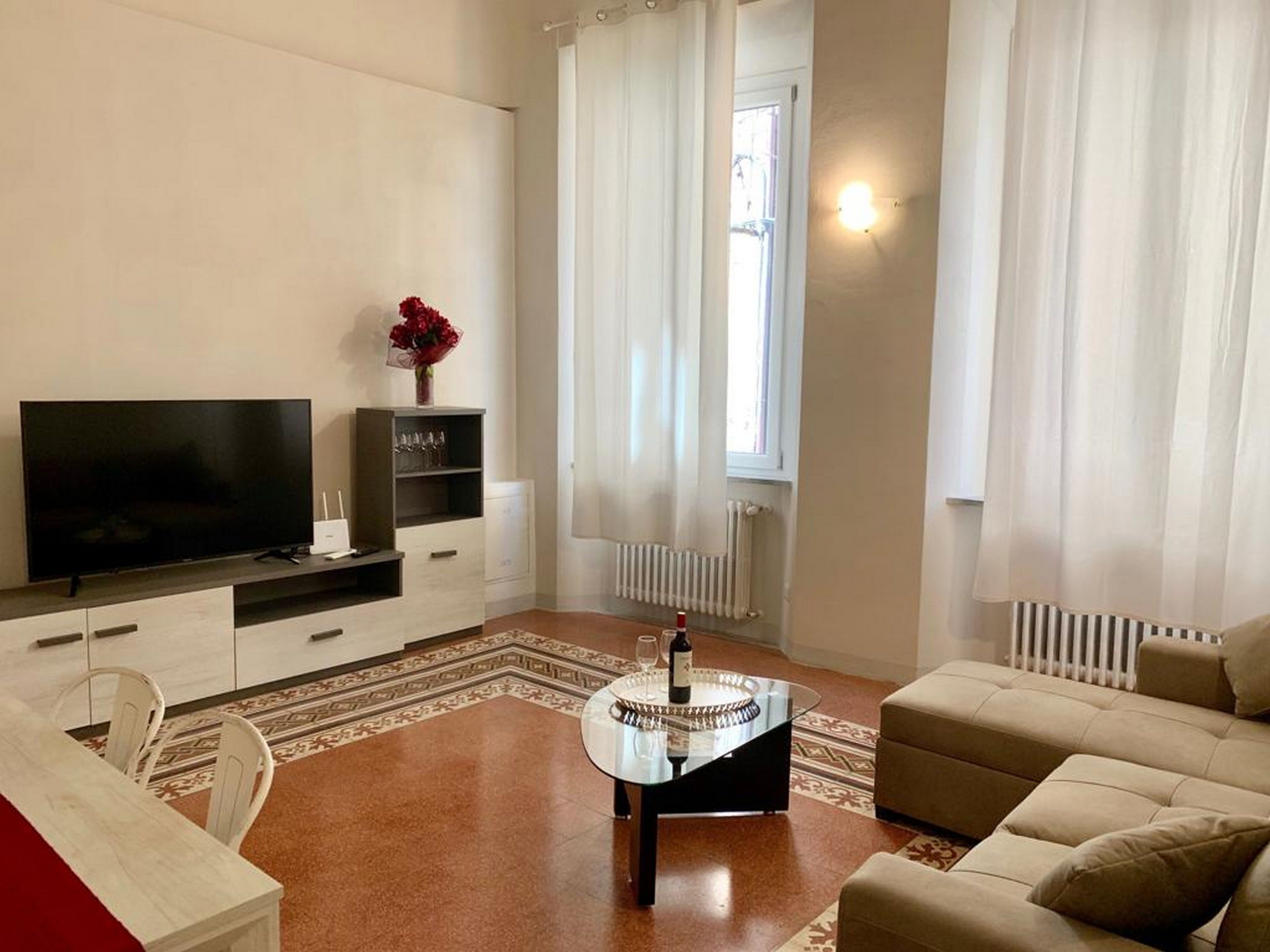 Uma sala de estar moderna com televisão de tela plana, mesa de centro de vidro, sofá bege e tapete estampado.