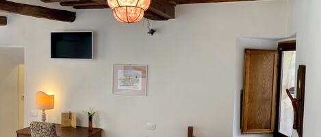 Ein Zimmer mit Holzboden, einem Schreibtisch mit Stuhl, einem Wand-Fernseher, einem gerahmten Bild und einem Fenster mit Holztür.