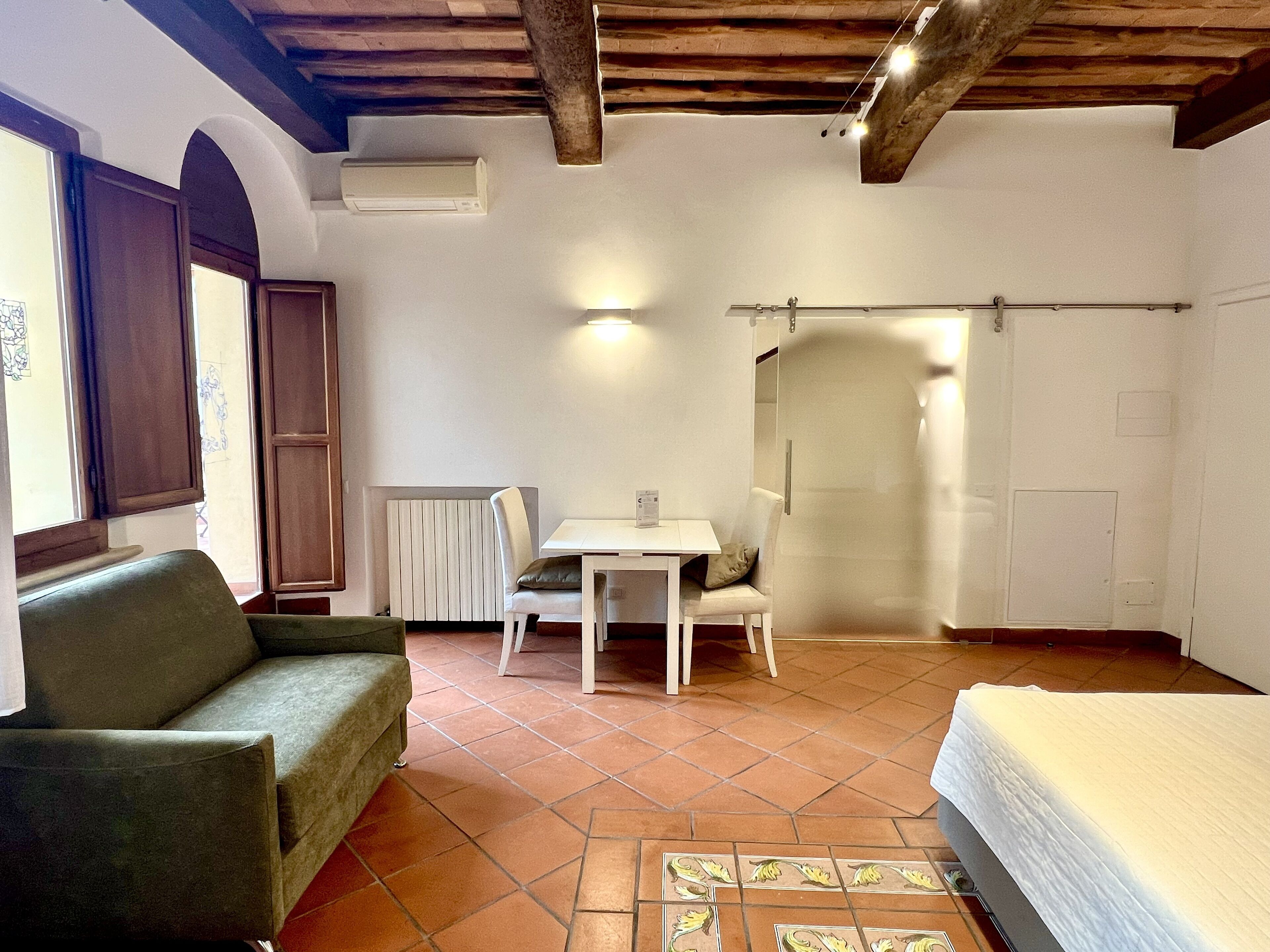 Una stanza con un divano verde, un tavolo bianco con una sedia, un letto, un termosifone e un soffitto in legno.