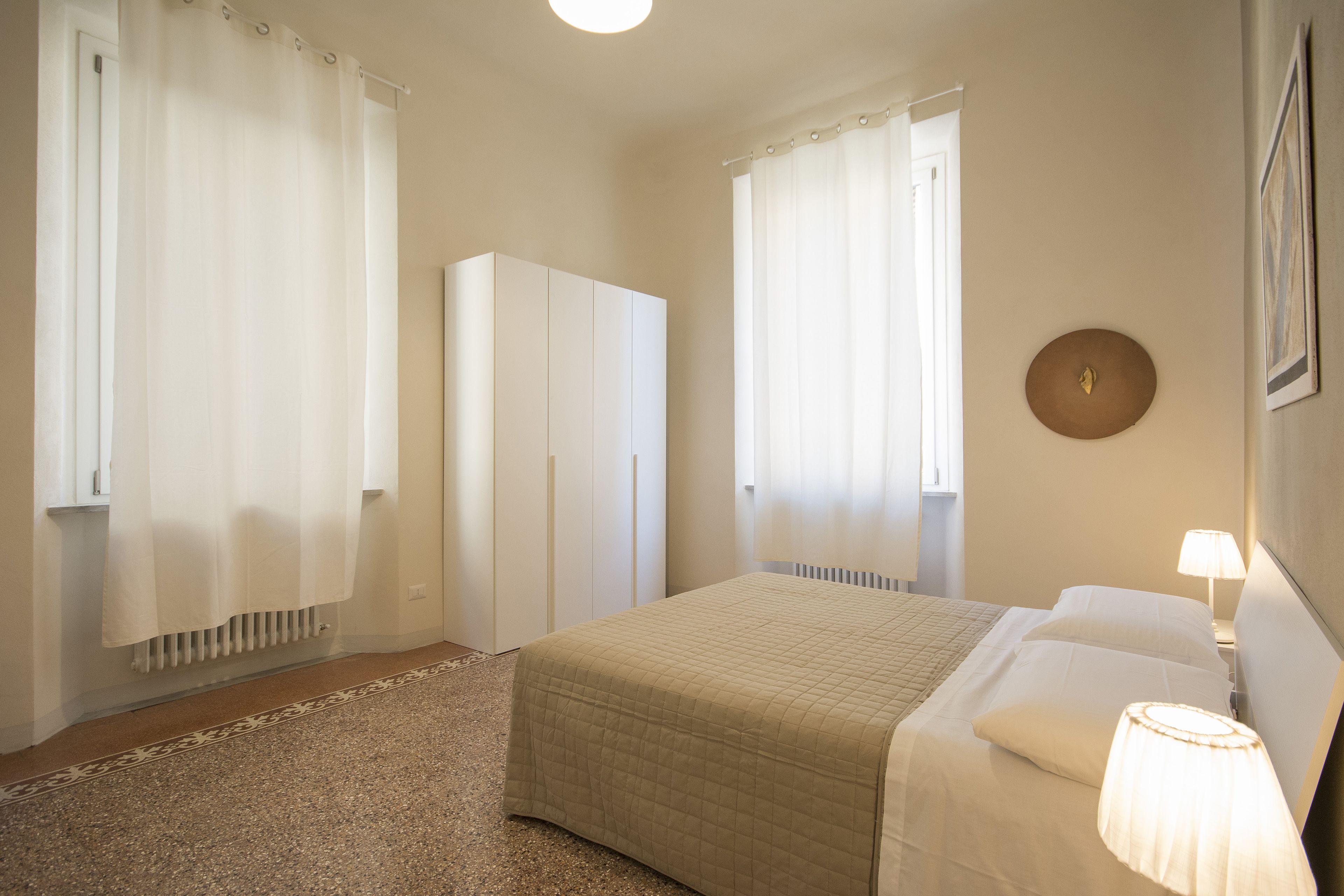 A bedroom with a bed, a nightstand, a lamp, a wardrobe, and a window with curtains.
