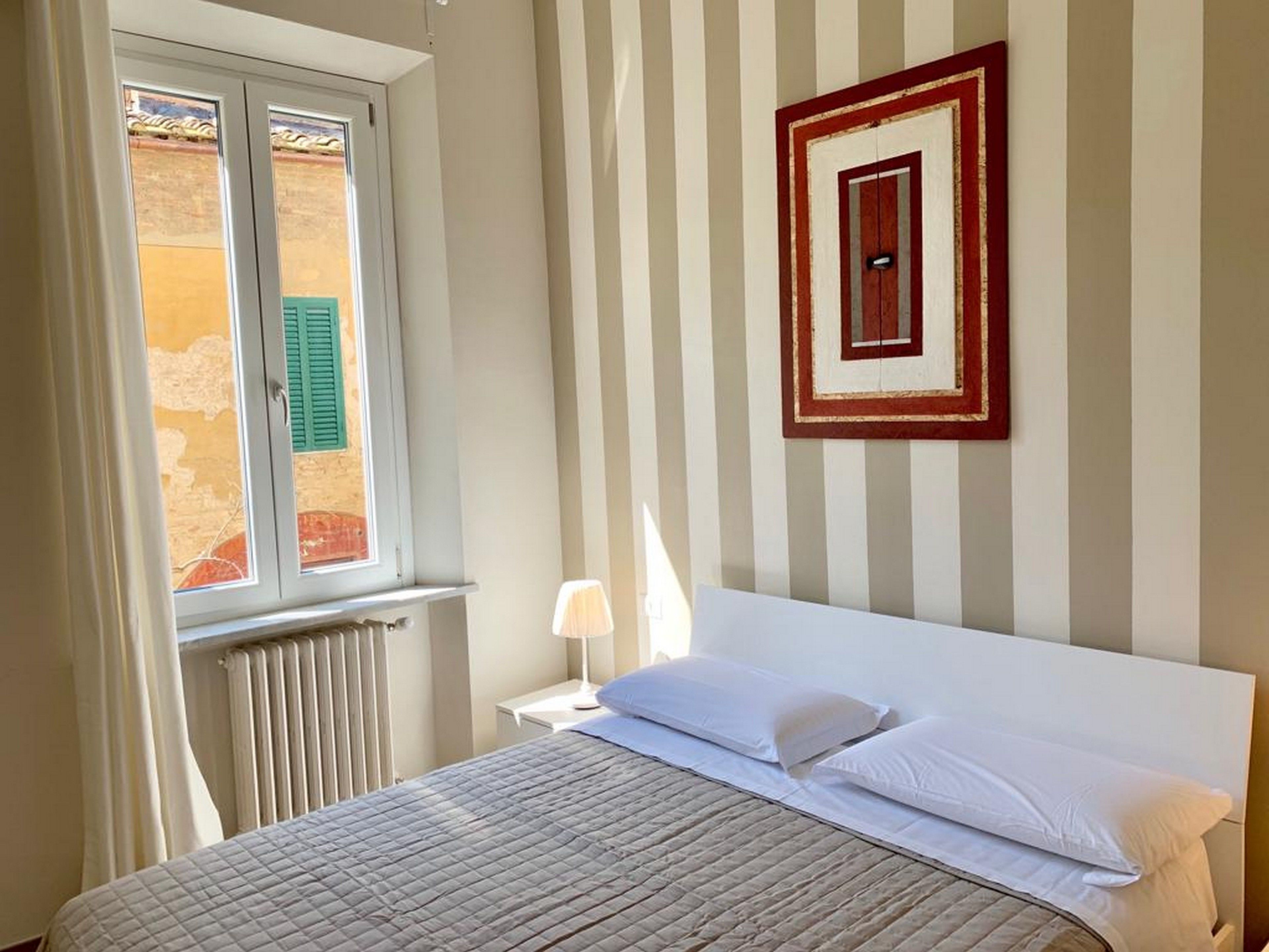 Una cama con ropa de cama blanca, una ventana con contraventanas verdes y una pared con rayas verticales y un cuadro enmarcado.