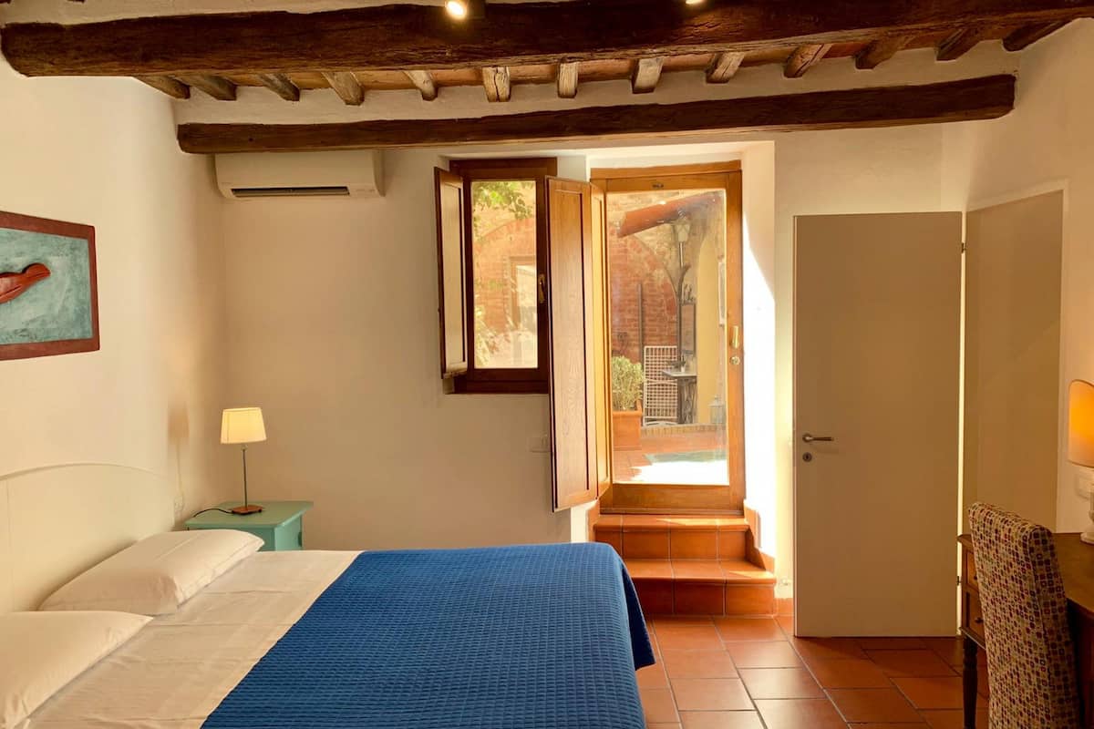 A bedroom with a bed, a nightstand, a lamp, a door leading to a balcony, and a view of the outside.