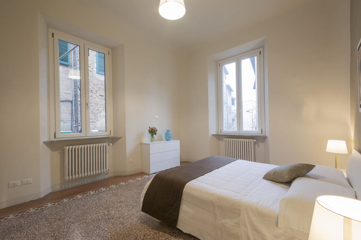 A bedroom with a bed, two windows, a radiator, a dresser, and a lamp.
