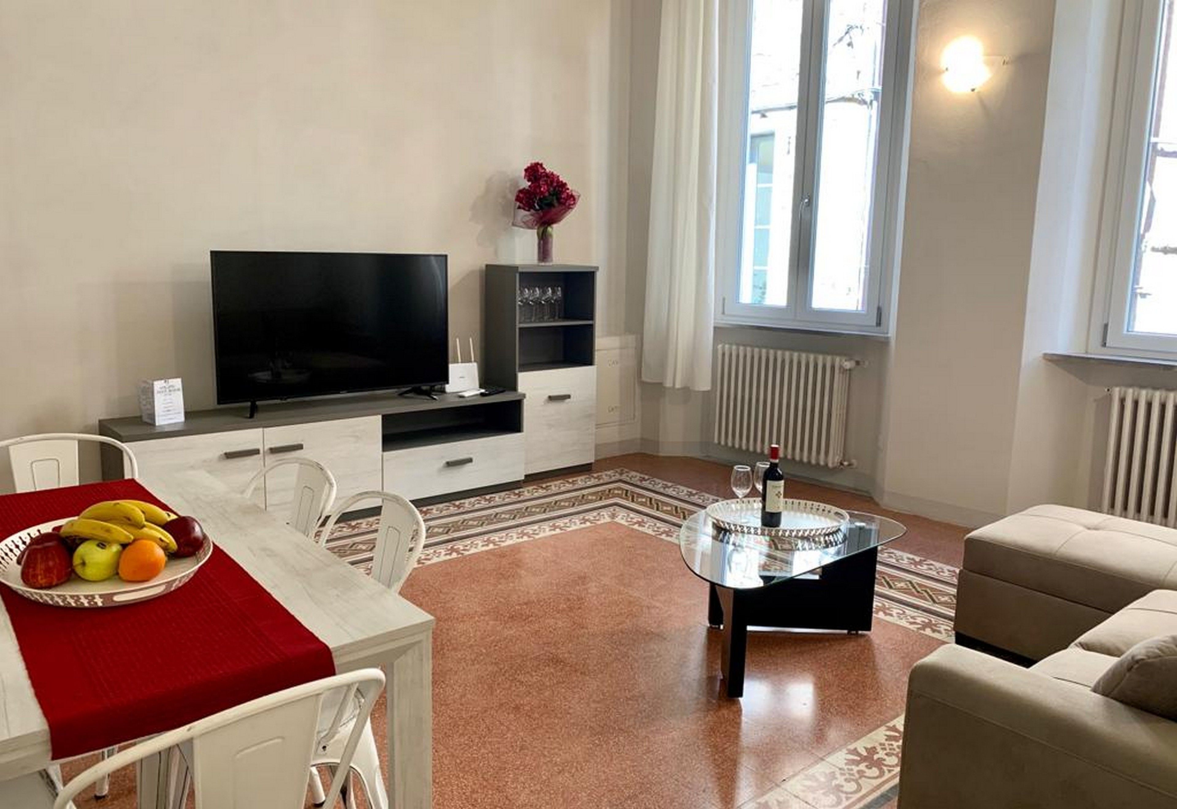 Un soggiorno moderno con una TV a schermo piatto, un tavolo da pranzo con frutta, un tavolino da caffè in vetro e un divano color beige.