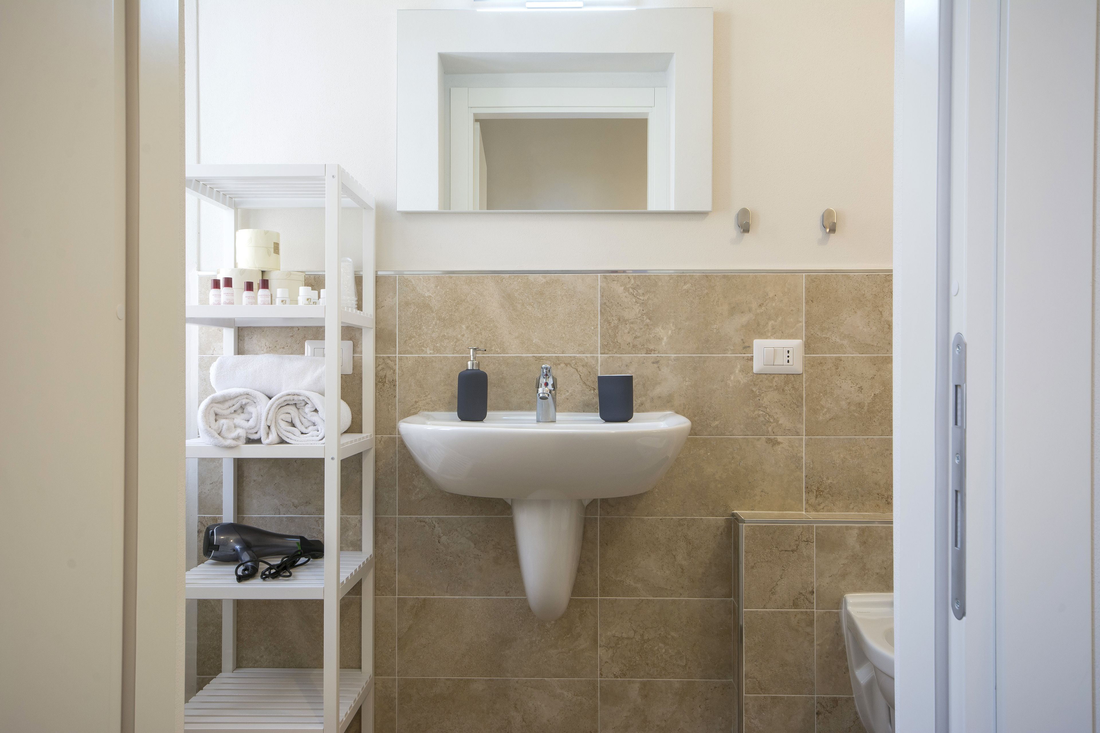 A bathroom with a white sink, beige tiles, a mirror above the sink, and shelves with towels and toiletries.