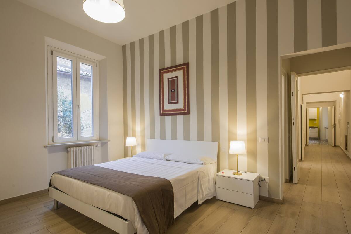 A bedroom with a bed, bedside tables, a window, a radiator, and a hallway leading to another room.