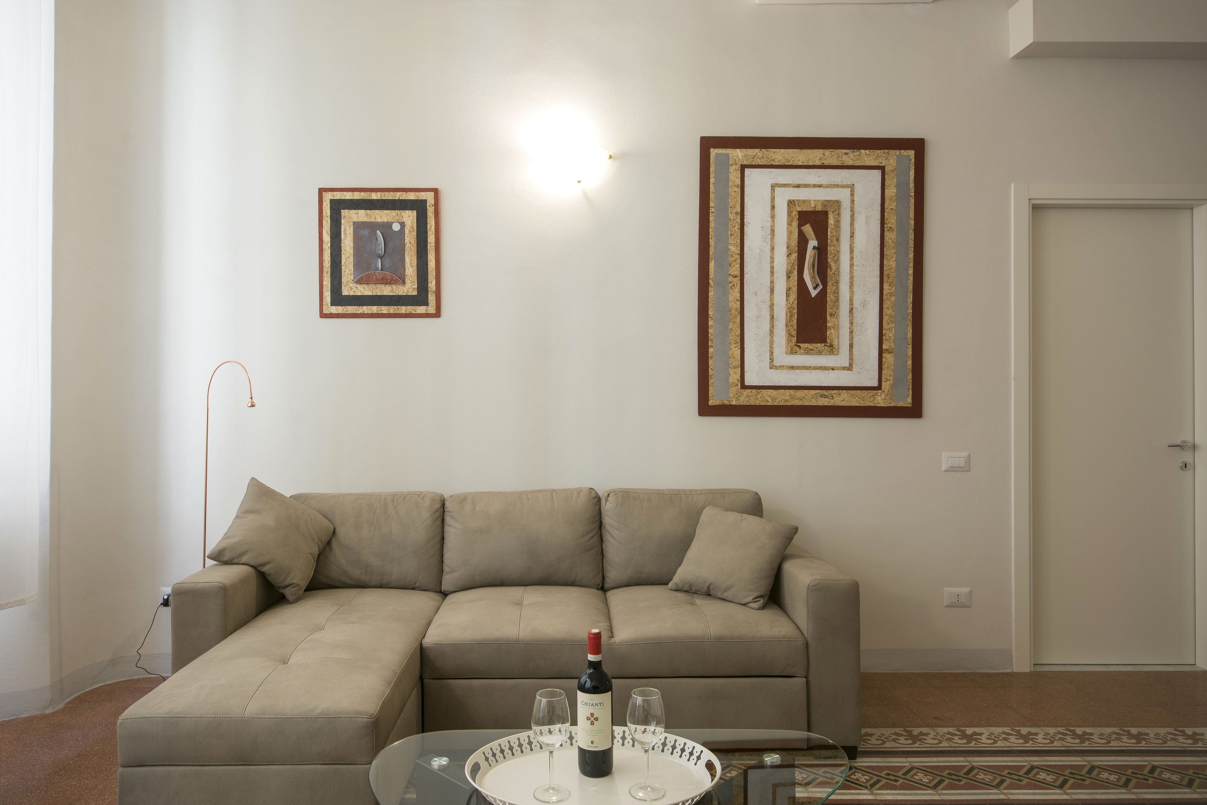 A living room with a beige sectional sofa, a glass coffee table with a wine bottle and two glasses, and two framed artworks on the wall.