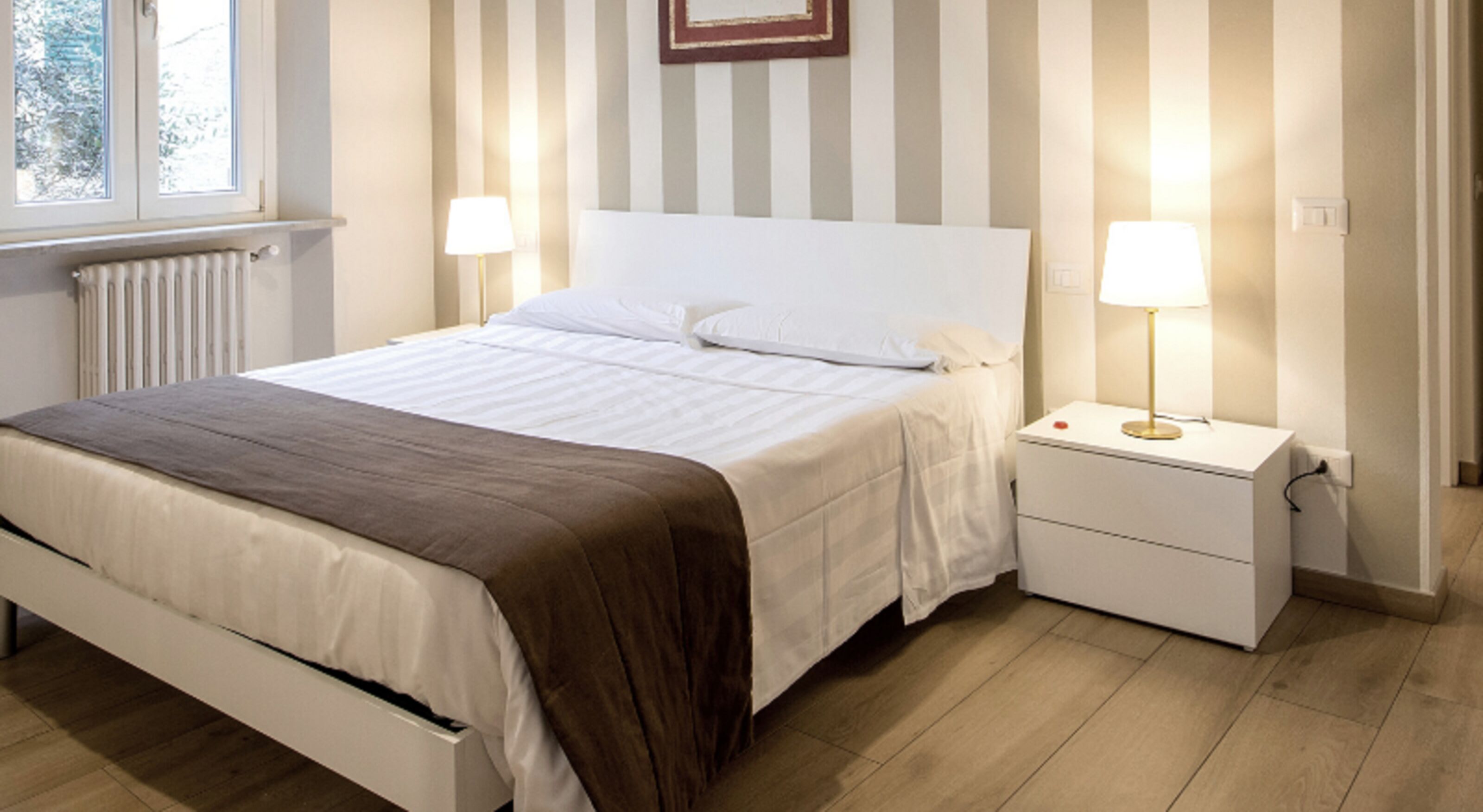 Une chambre à coucher avec un lit, des tables de chevet, une lampe, un radiateur et une fenêtre.