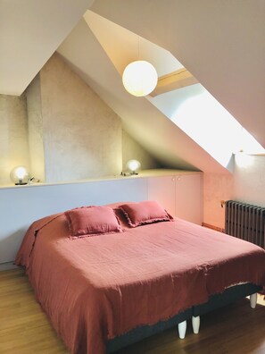 Une chambre avec un plafond en pente, un lit avec une literie rose et deux lampes fixées au mur.