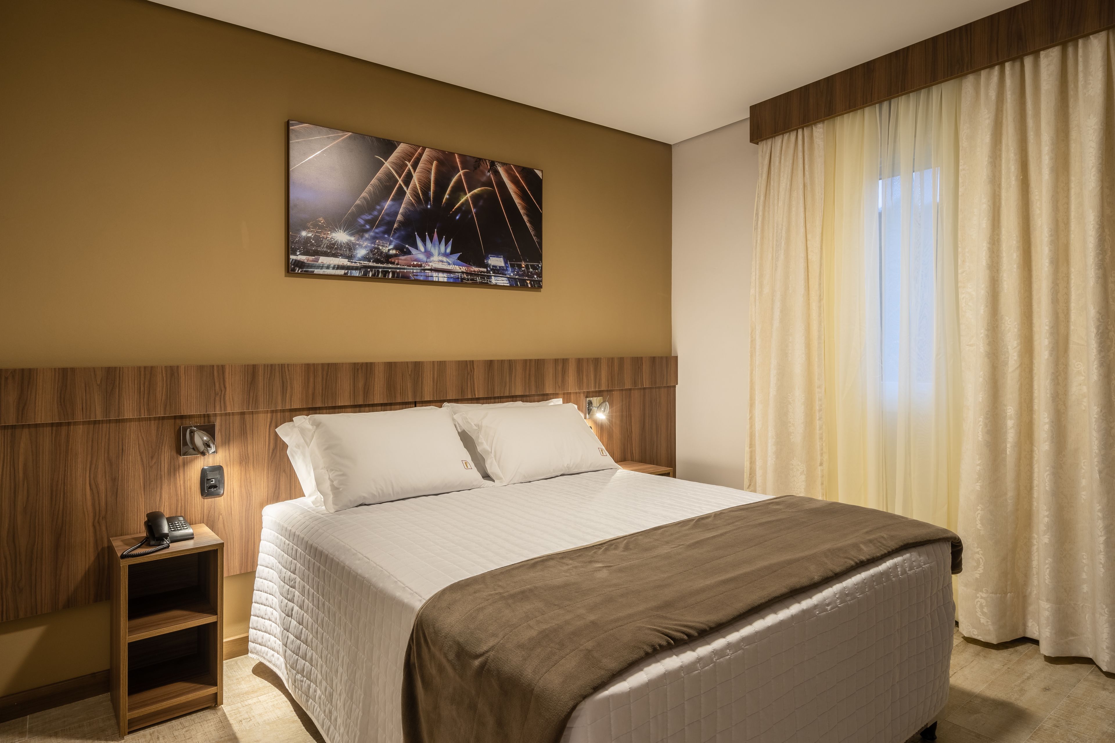 A hotel room with a large bed, a nightstand with a phone, a wall-mounted TV, and a window with curtains.