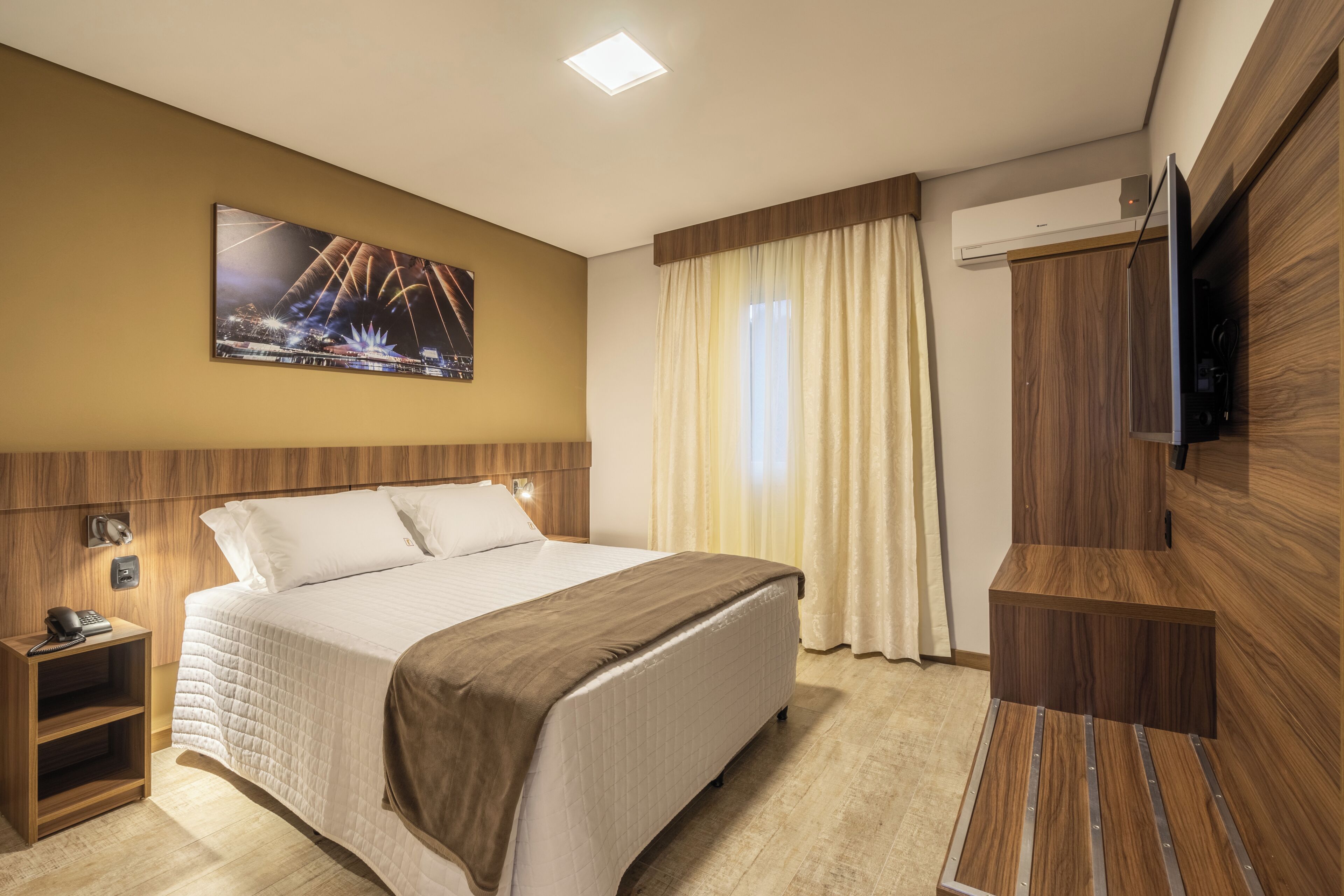 Ein Hotelzimmer mit Bett, Fernseher, Nachttisch, Wandklimaanlage und einem Vorhangfenster.