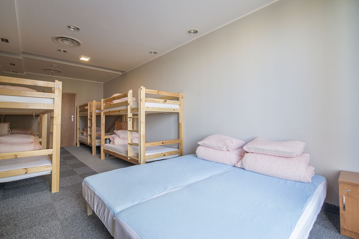 Quarto com beliches e uma cama simples, com iluminação no teto e um pequeno armário de madeira.
