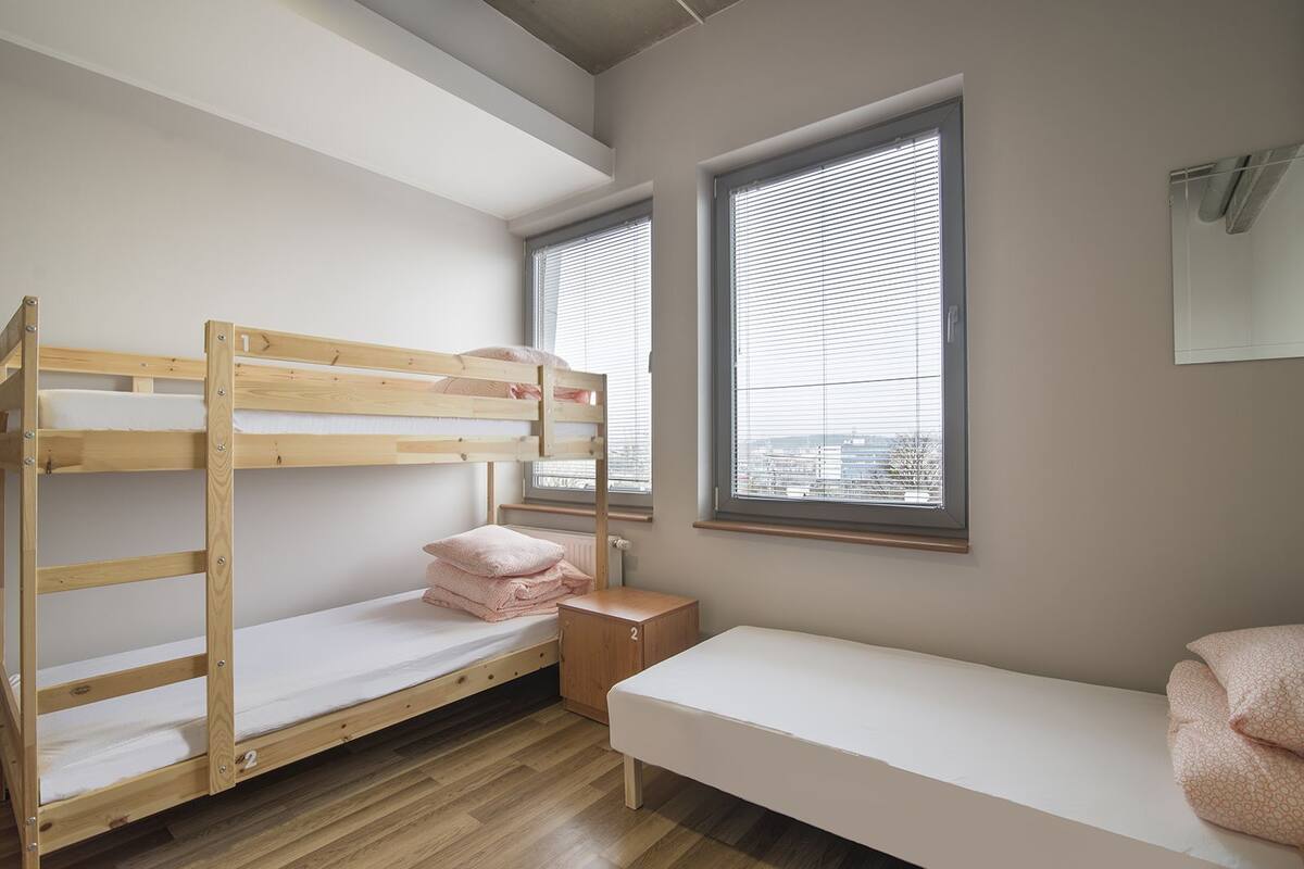 Quarto com beliche, uma pequena mesa de cabeceira de madeira, uma cama de solteiro e duas janelas com persianas.