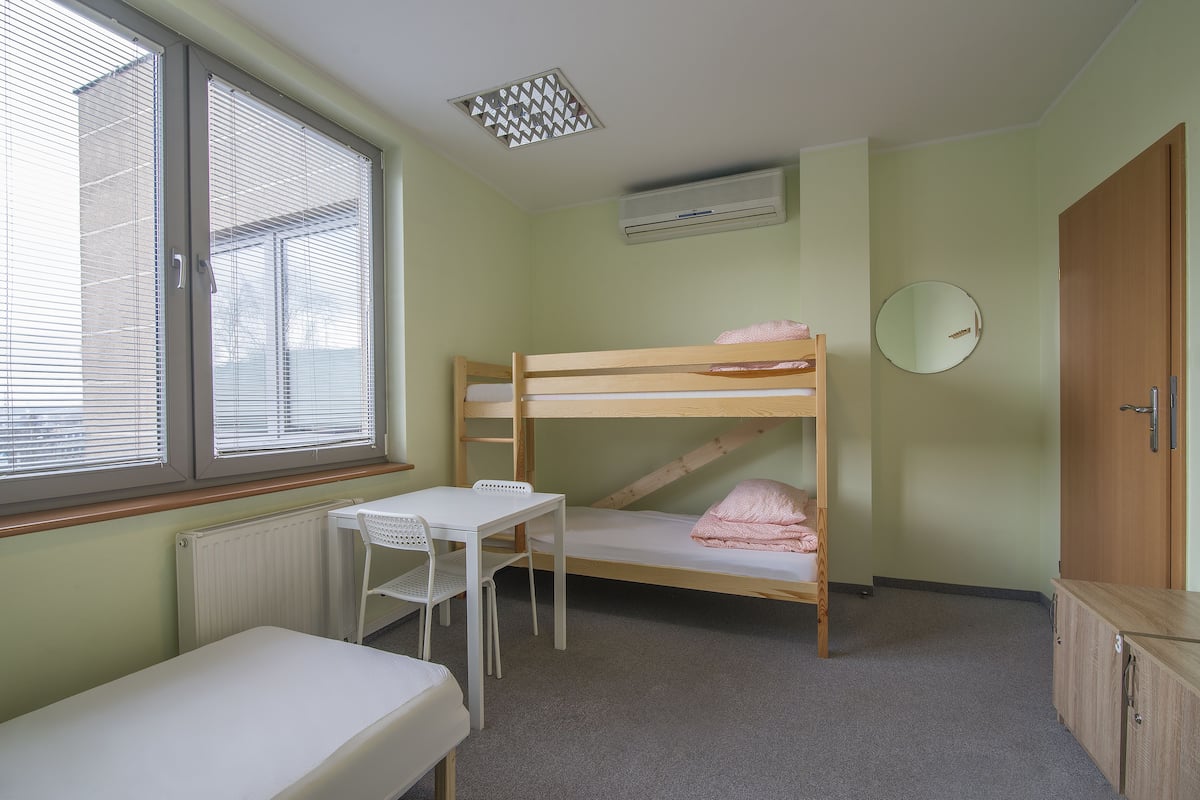 Quarto com uma cama beliche, uma mesa pequena, uma cadeira, um radiador, uma janela com persianas, um aparelho de ar condicionado e um guarda-roupa de madeira.