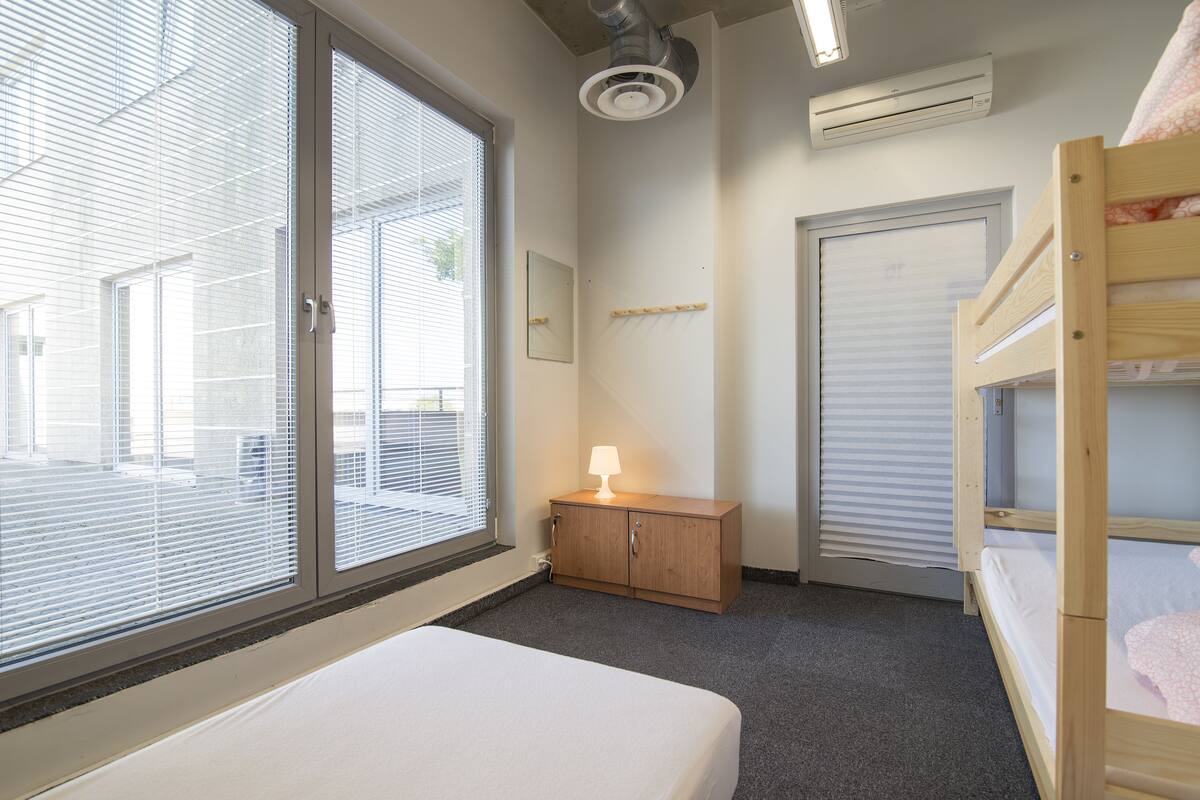 Quarto com uma cama de solteiro, mesa de cabeceira com abajur, janela com persianas e ar condicionado.
