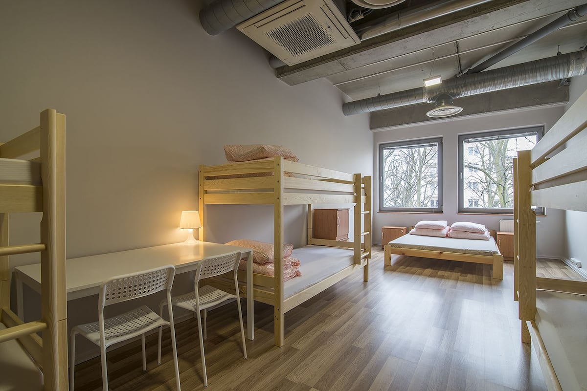 Quarto com beliche, escrivaninha e cadeiras, janela com vista para árvores e teto com dutos aparentes.