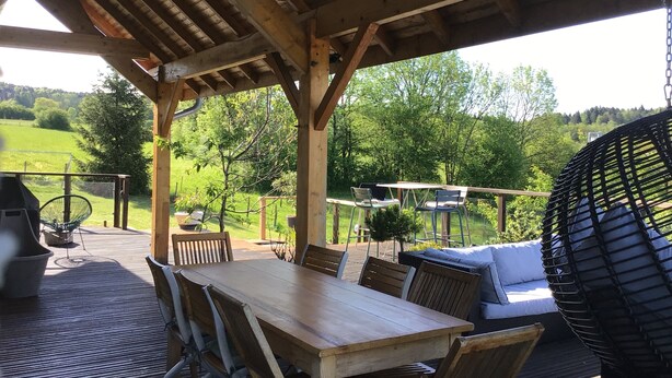 An outdoor wooden deck with a table and chairs, surrounded by lush greenery and trees. - 220 m² house, sleeps 8–10. With private grounds and a terrace overlooking the lake and spa (Hautecour)