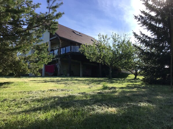 A two-story building with a sloped roof, surrounded by trees and a grassy yard. - 220 m² house, sleeps 8–10. With private grounds and a terrace overlooking the lake and spa (Hautecour)