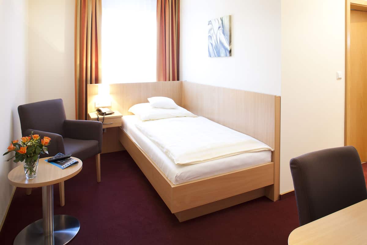 A hotel room with a single bed, a desk with a chair, a small table with a vase of flowers, and a nightstand with a lamp and phone.
