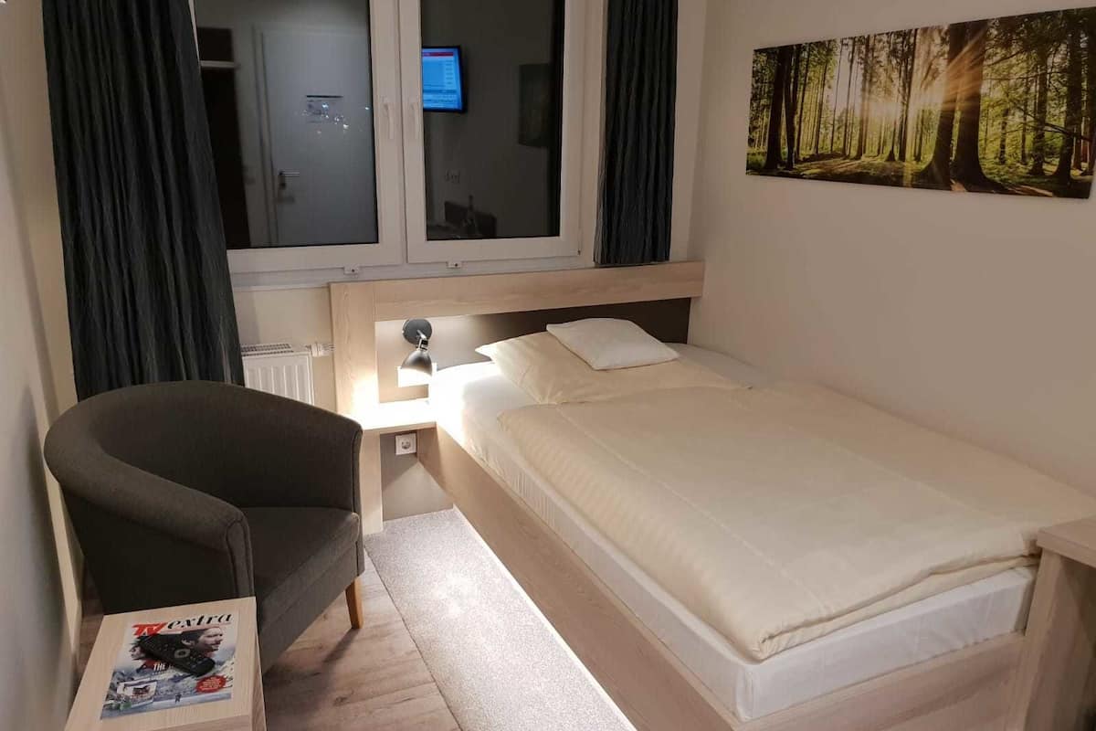 A bedroom with a bed, a chair, a nightstand, a TV, and a forest painting on the wall.