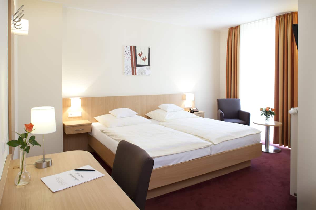 A hotel room with a double bed, bedside tables with lamps, a desk with a chair, a vase with a flower, and a small table with a vase of flowers.