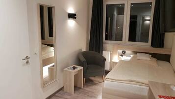 Habitación de hotel con cama, una silla, una mesita, un espejo y una ventana con cortinas.