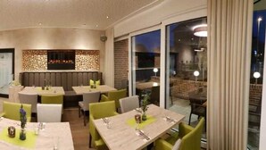 A modern restaurant with booths and tables, a bar area, and a view of the cityscape. - Hotel Clemenswerther Hof (Sögel)