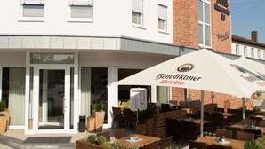 A hotel with a white facade, a brick section, outdoor seating with umbrellas, and a sign for 'Benediktiner Weissbier'. - Hotel Clemenswerther Hof (Sögel)