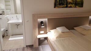 A bedroom with a wooden headboard, a bedside table with a lamp, a bed with pillows, a painting of a forest on the wall, and a bathroom with a sink and mirror. - Hotel Clemenswerther Hof (Sögel)