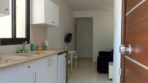 A compact kitchen with white cabinets, a sink, and a small refrigerator. There is a dining area with a chair and a purple sofa. - Royal Sunset Studios (Limassol)
