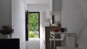 A narrow hallway leading to a kitchen with a white table and chairs, a potted plant, and a view of greenery outside. - Royal Sunset Studios (Limassol)