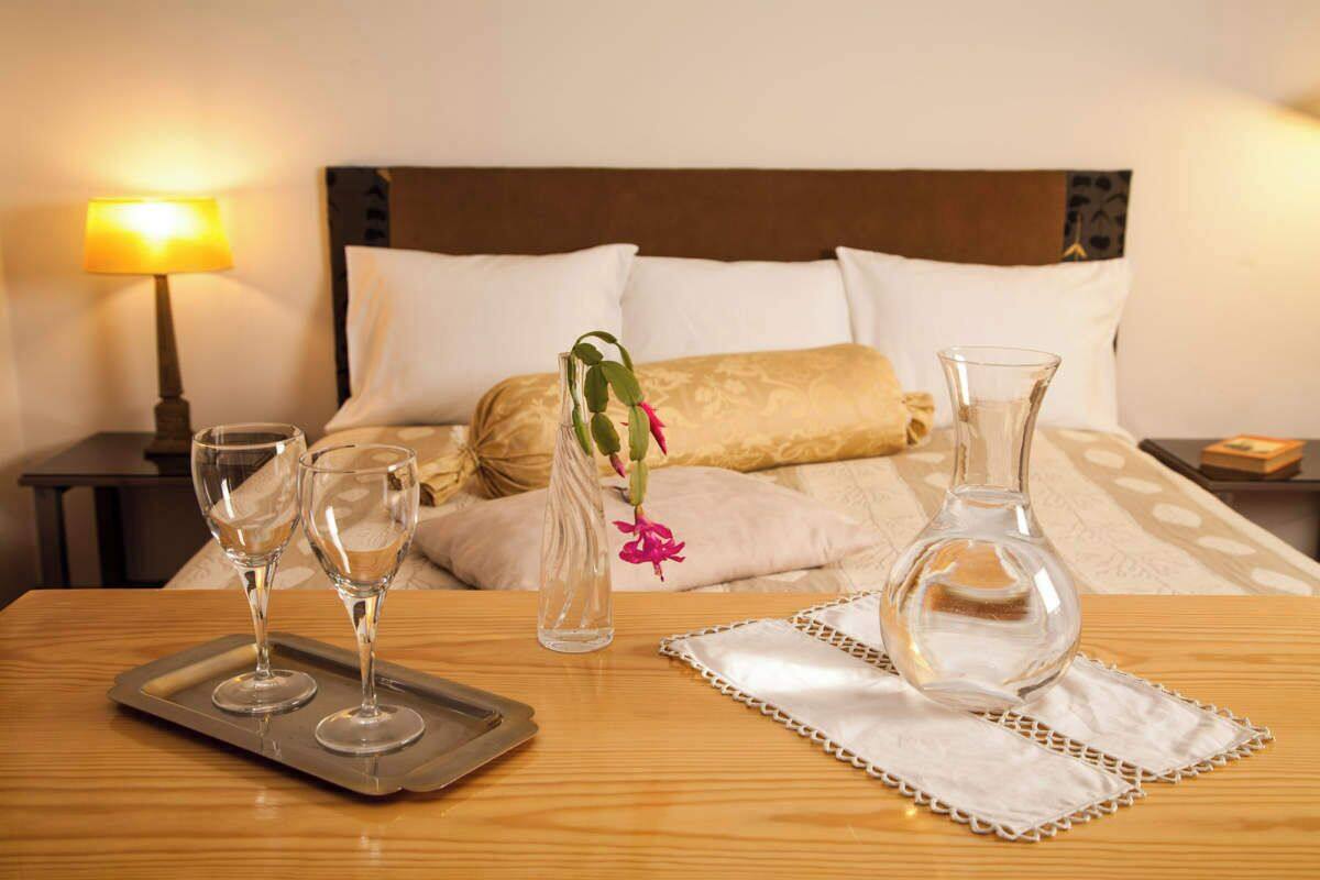 A neatly arranged hotel room with a bed, a bedside table with a lamp, and a tray with two glasses and a vase with flowers.