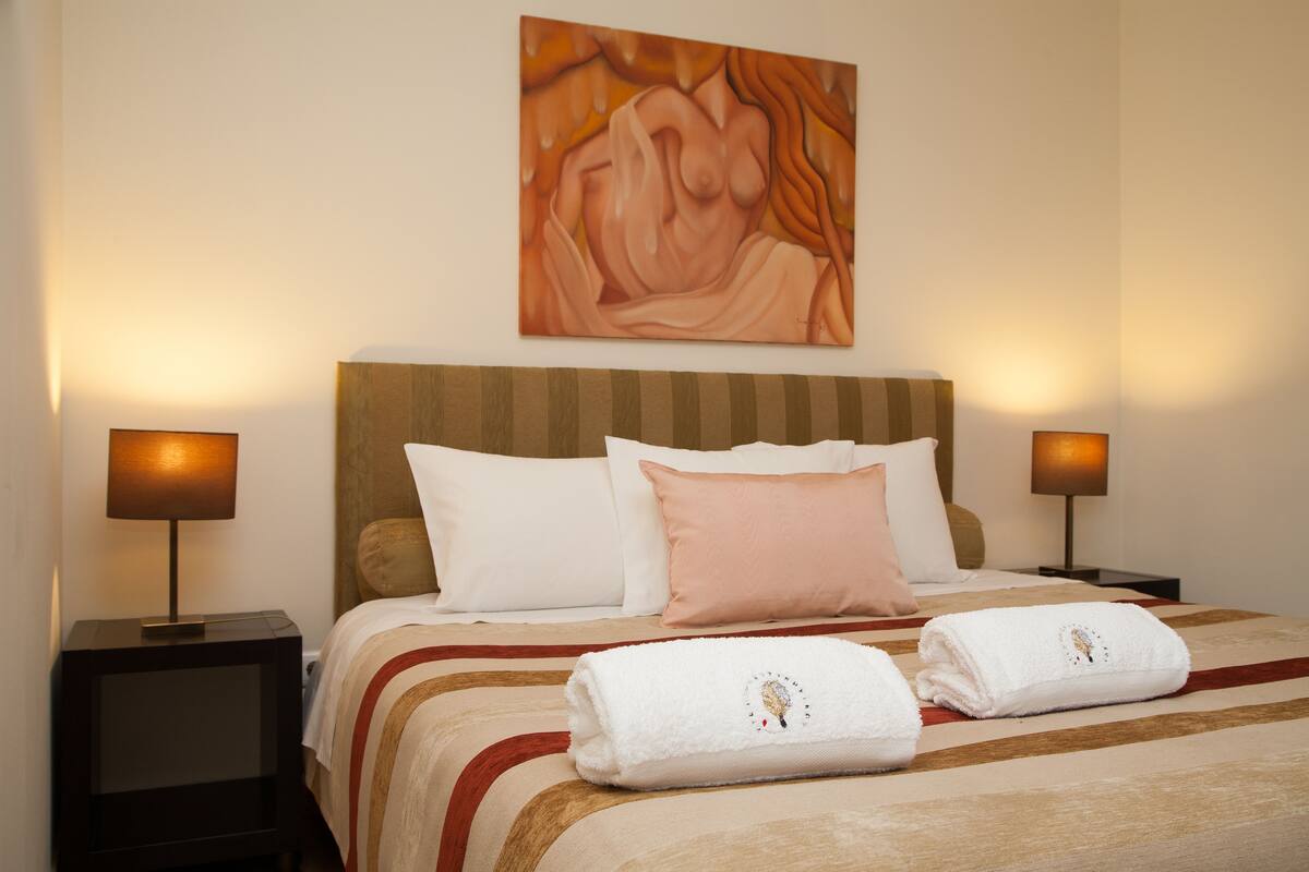 A hotel room with a large bed, two bedside lamps, a painting on the wall, and two towels on the bed.