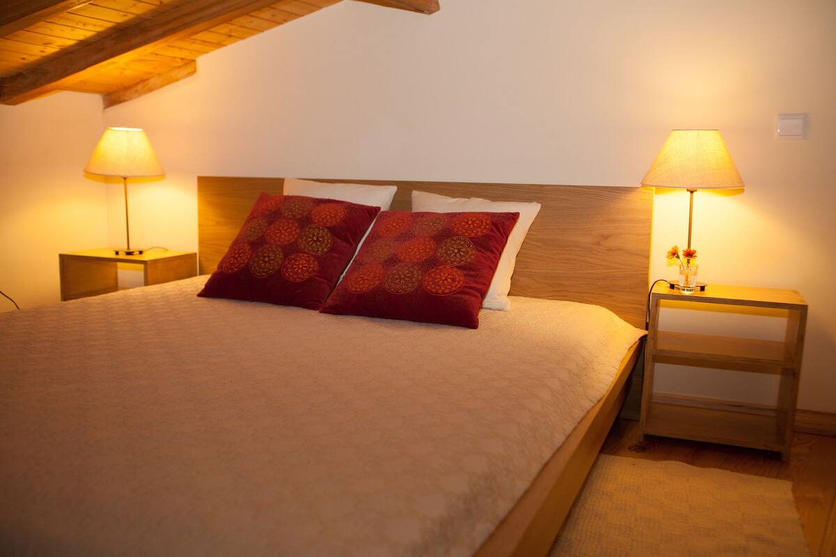 A neatly made bed with two red pillows, two bedside lamps, and a wooden nightstand with a vase of flowers.
