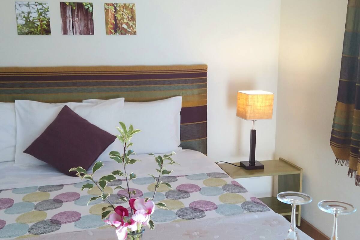 A hotel room with a bed, a bedside table with a lamp, a vase with flowers, and a table with a napkin and glasses.