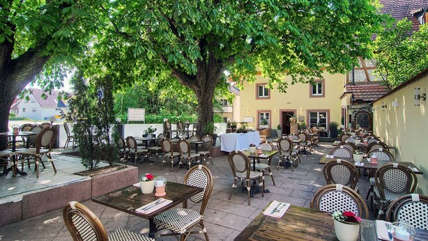 An outdoor café with wooden tables and wicker chairs, surrounded by trees and buildings.