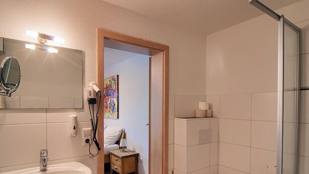Een badkamer met een wastafel, toilet en douche. Een deur leidt naar een slaapkamer met een bed en nachtkastje.