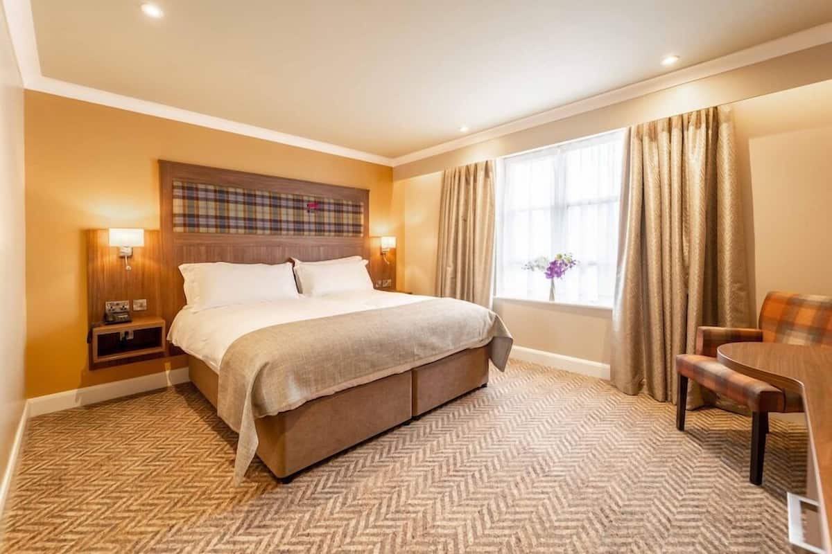 Quarto de hotel com cama grande, mesa de cabeceira, cadeira, mesinha e janela com cortinas.