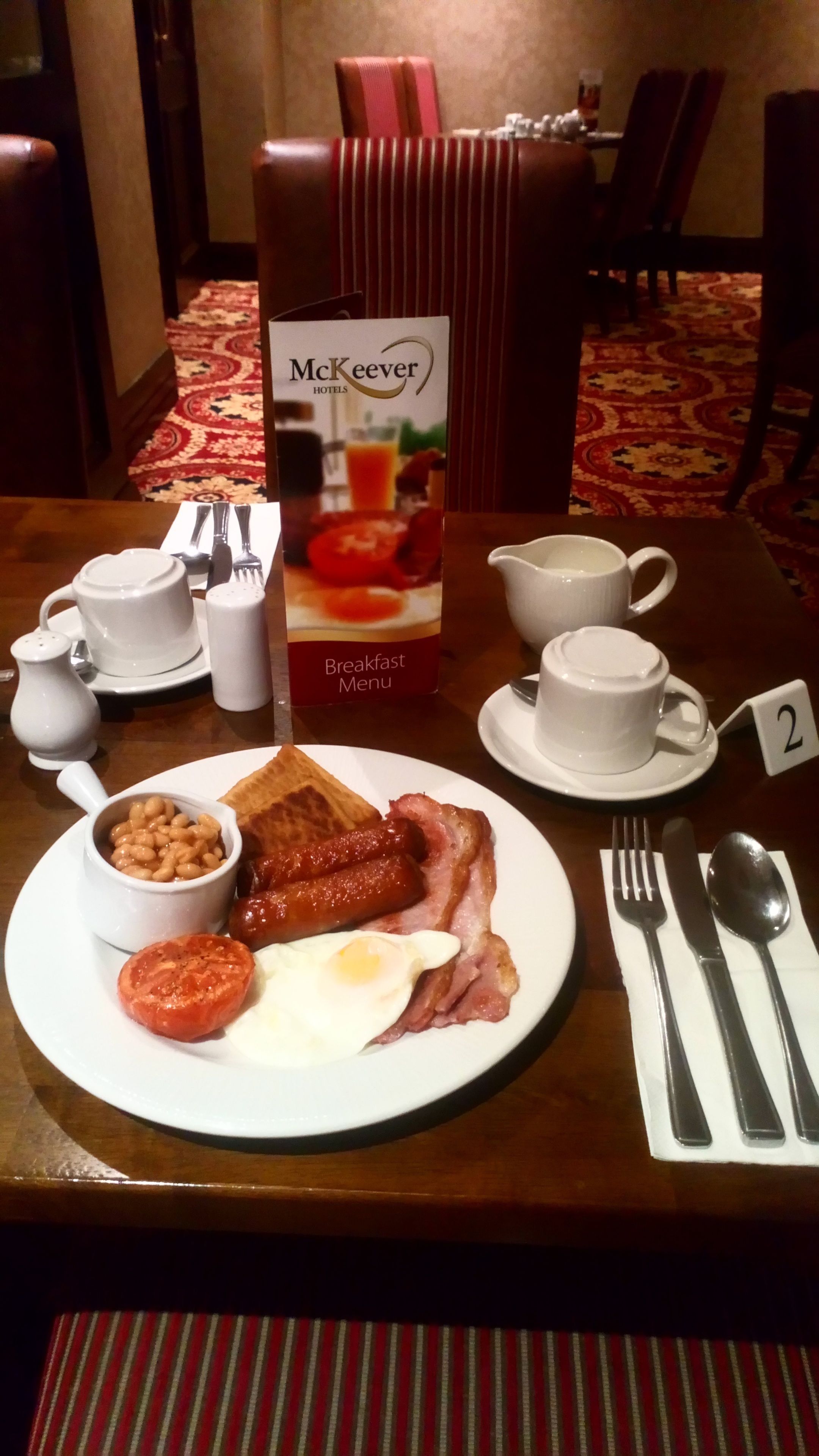 A breakfast table set with a plate of eggs, bacon, beans, and tomato, a coffee cup, and a breakfast menu.