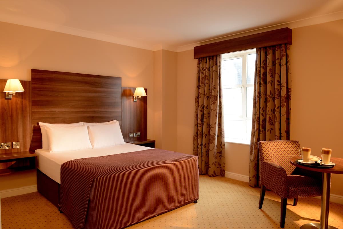 Quarto de hotel com uma cama grande, duas luminárias de cabeceira, uma cadeira, uma mesinha com duas chávenas e uma janela com cortinas.