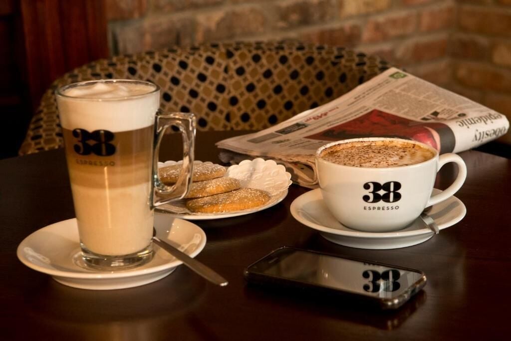 A table with a coffee cup labeled "38 ESPRESSO," a tall glass of coffee, a plate with cookies, and a newspaper.