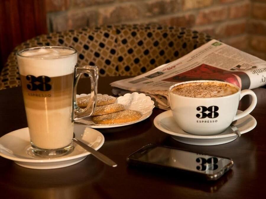 A table with a coffee cup labeled "38 ESPRESSO," a tall glass of coffee, a plate with cookies, and a newspaper.